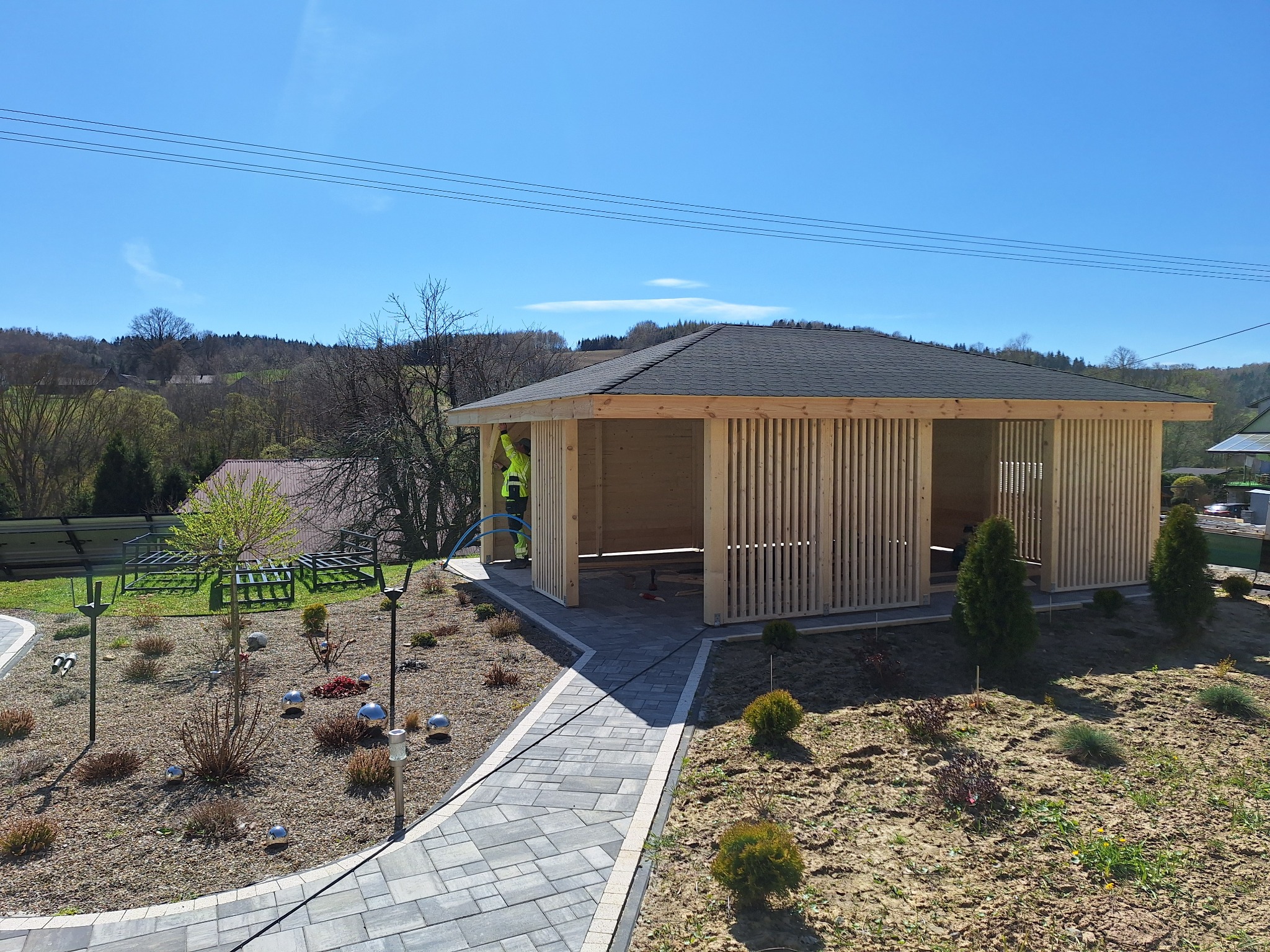 Nowoczesna altana z drewnianymi lamelami i ciemnym dachem na tle zieleni. Płytkowy chodnik prowadzi do wejścia. Widoczny fragment osoby w odblaskowej kamizelce.