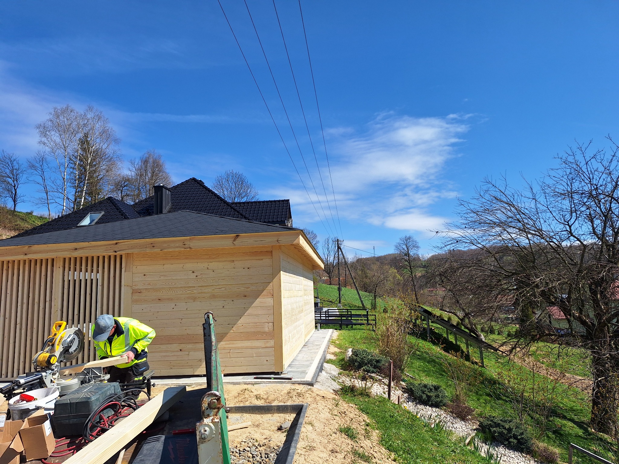 Pracownik w odblaskowej kamizelce tnie drewno piłą mechaniczną na tle drewnianej altany z czarnym dachem i błękitnego nieba. Widoczne narzędzia i otoczenie w trakcie budowy.