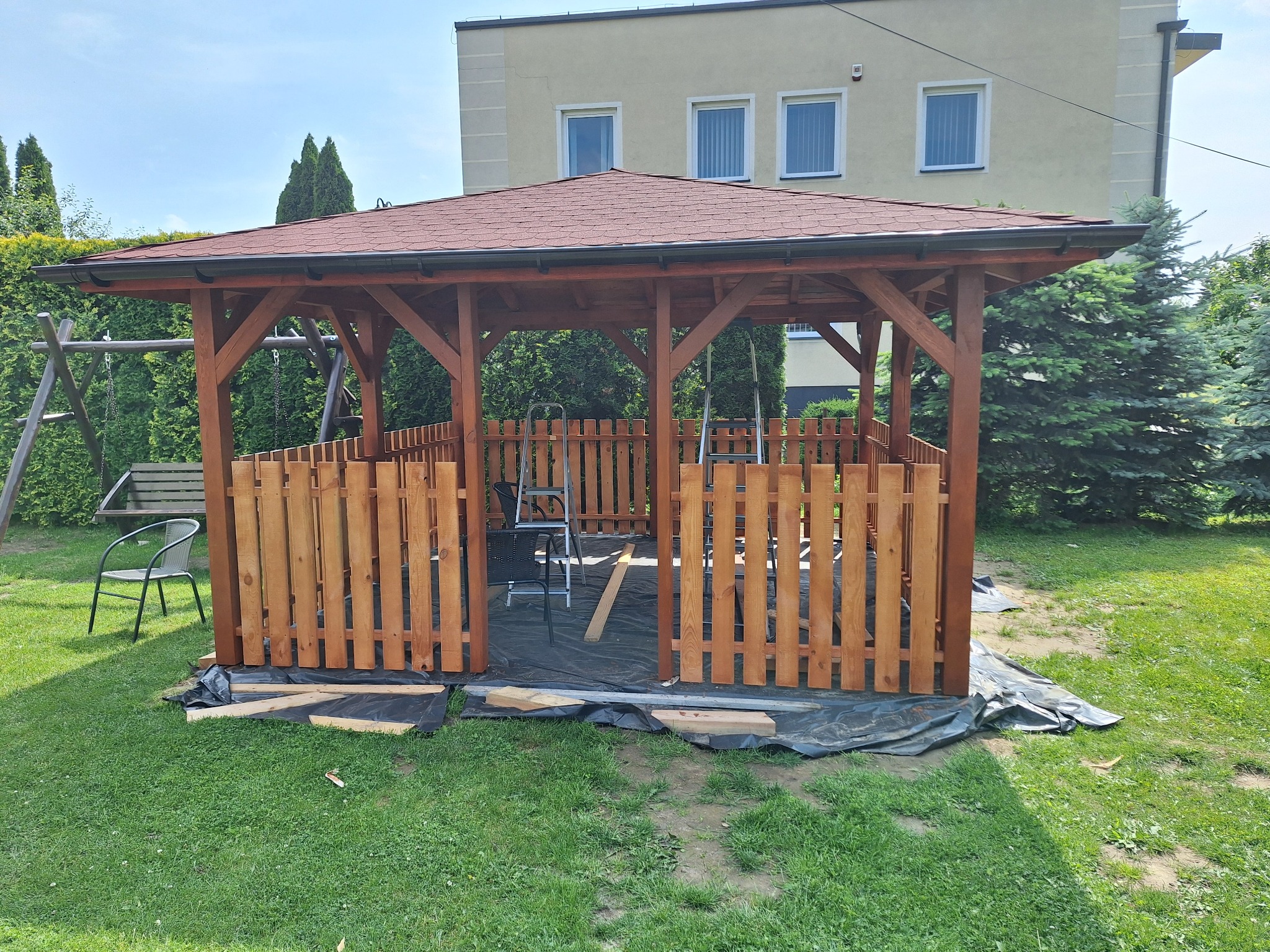 Drewniana altana z brązowym dachem, częściowo obudowana płotkiem, w trakcie budowy na trawniku. Widoczne narzędzia i materiały budowlane.