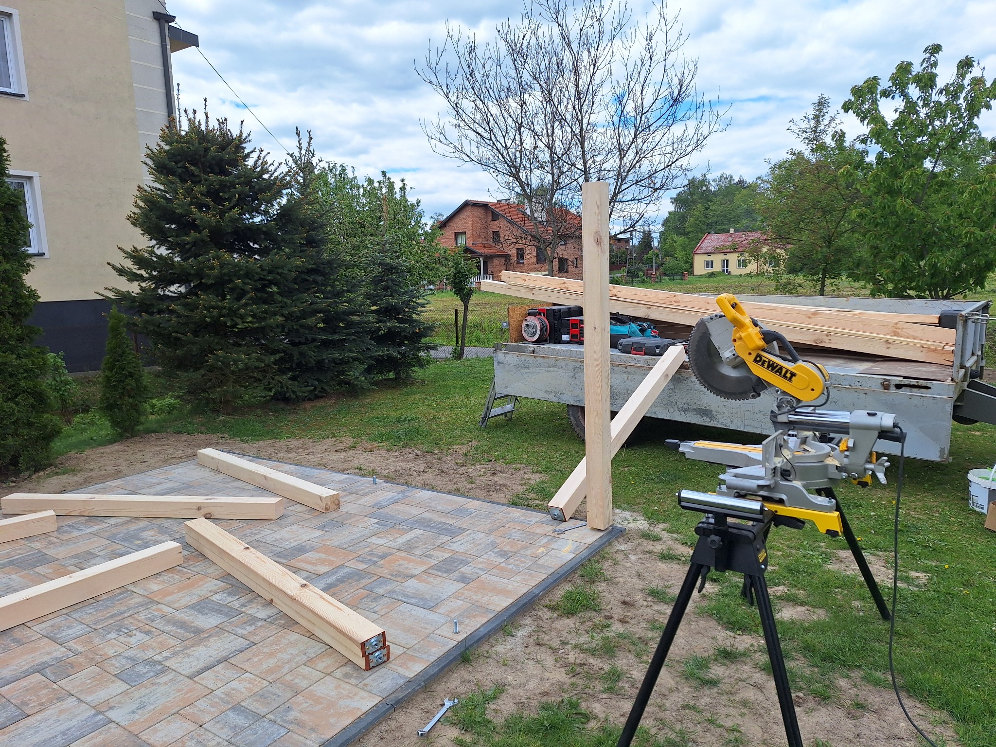 Budowa altany: przygotowane drewniane elementy na kostce, piła ukośnica DeWalt, przyczepka z drewnem w tle, domy i drzewa w tle. Widok z poziomu gruntu.