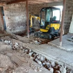 Żółta minikoparka Yanmar w trakcie prac ziemnych wewnątrz budynku o ceglanych ścianach i betonowym stropie, widoczny wykop wypełniony gruzem i ślady opon.