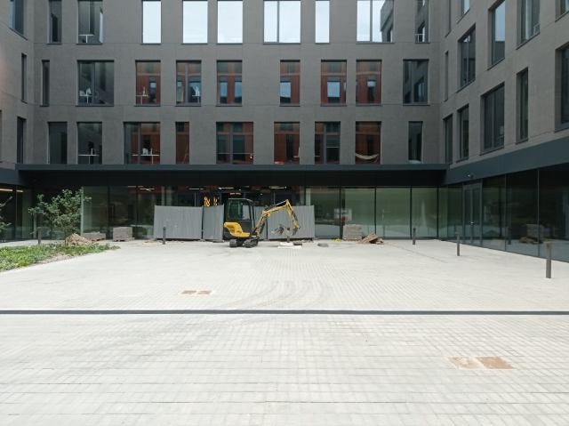 Żółta mini koparka stoi na placu budowy przed nowoczesnym budynkiem z licznymi oknami, widoczny fragment wykopu zabezpieczony szarą blachą falistą, na placu leżą stosy kostki brukowej.