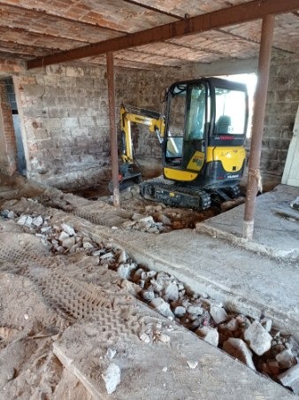 Żółta minikoparka Yanmar w trakcie prac ziemnych wewnątrz budynku o ceglanych ścianach i betonowym stropie, widoczny wykop wypełniony gruzem i ślady opon.