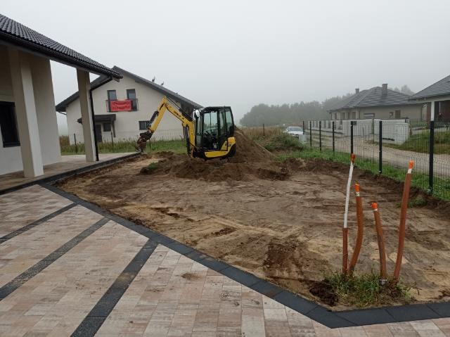 Żółta mini koparka na placu budowy w trakcie prac ziemnych, widoczne wystające rury z pomarańczowymi zaślepkami, kostka brukowa i domy w tle.