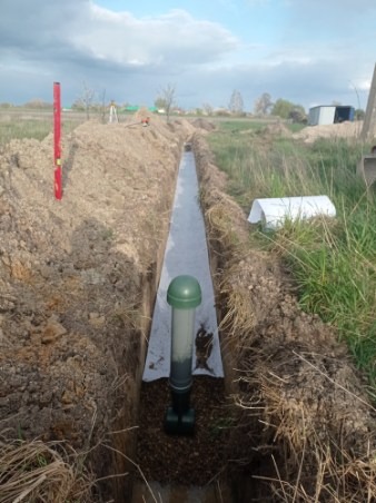 Długi wykop w ziemi wyłożony geowłókniną, z pionowo osadzonym drenem inspekcyjnym z zieloną pokrywą, w tle poziomica i niwelator optyczny.