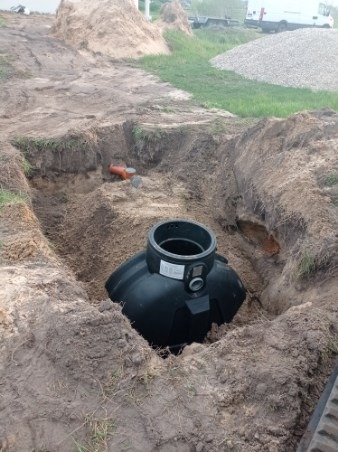 Czarny zbiornik z tworzywa sztucznego umieszczony w wykopie ziemnym, widoczny pomarańczowy rurociąg, w tle sterta ziemi i zaparkowany biały samochód dostawczy.