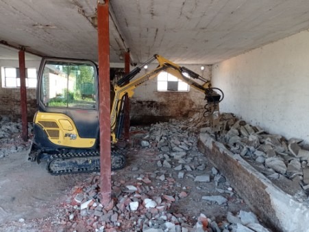 Żółto-czarna mini koparka Yanmar SV19 z hydraulicznym młotem wyburzeniowym kruszy gruz wewnątrz budynku z ceglanymi ścianami, oświetlonego naturalnym światłem z okna.