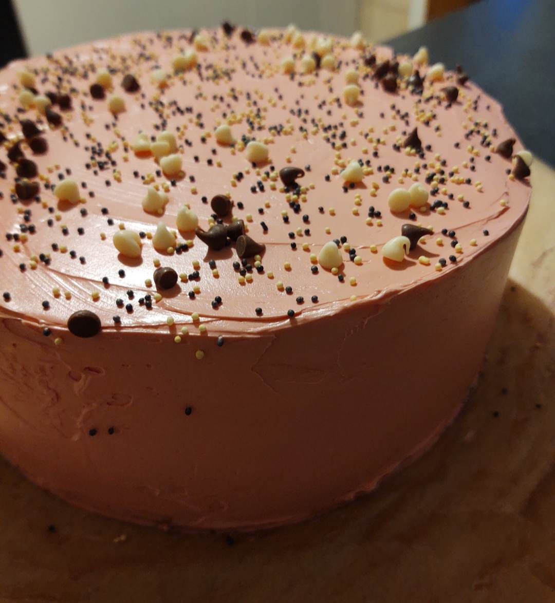 Różowy tort z czekoladowymi i białymi dropsami oraz kolorową posypką na drewnianej desce.