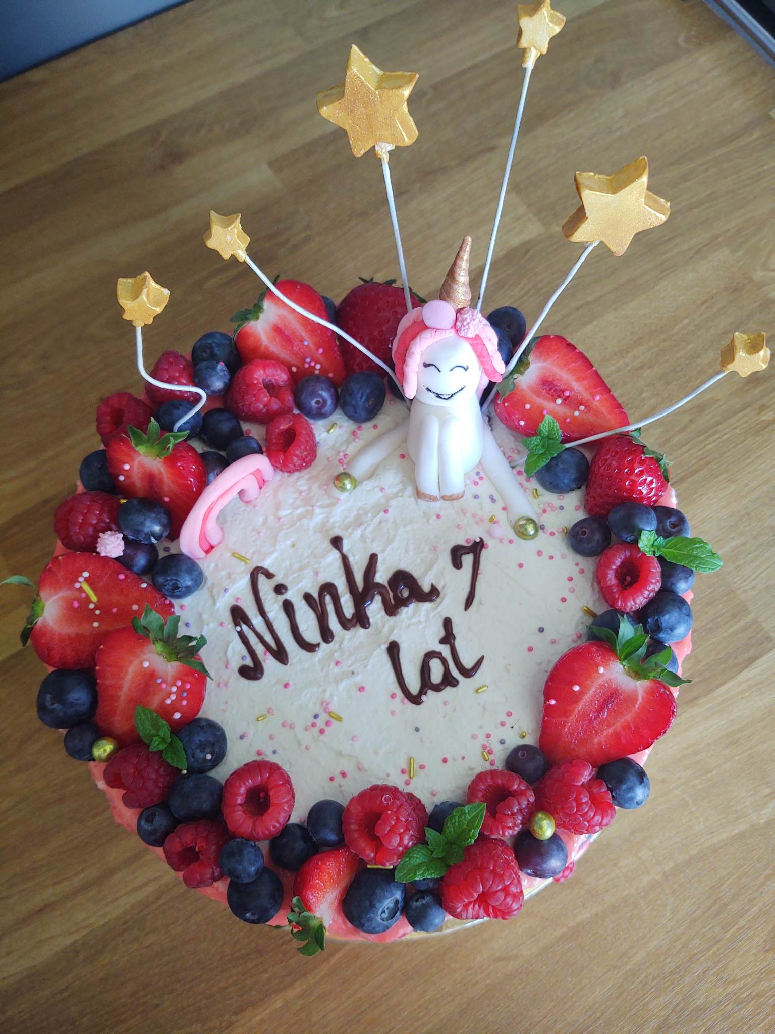 Tort urodzinowy z jednorożcem, owocami (truskawki, maliny, borówki) i napisem 'Ninka 7 lat' na drewnianym stole.