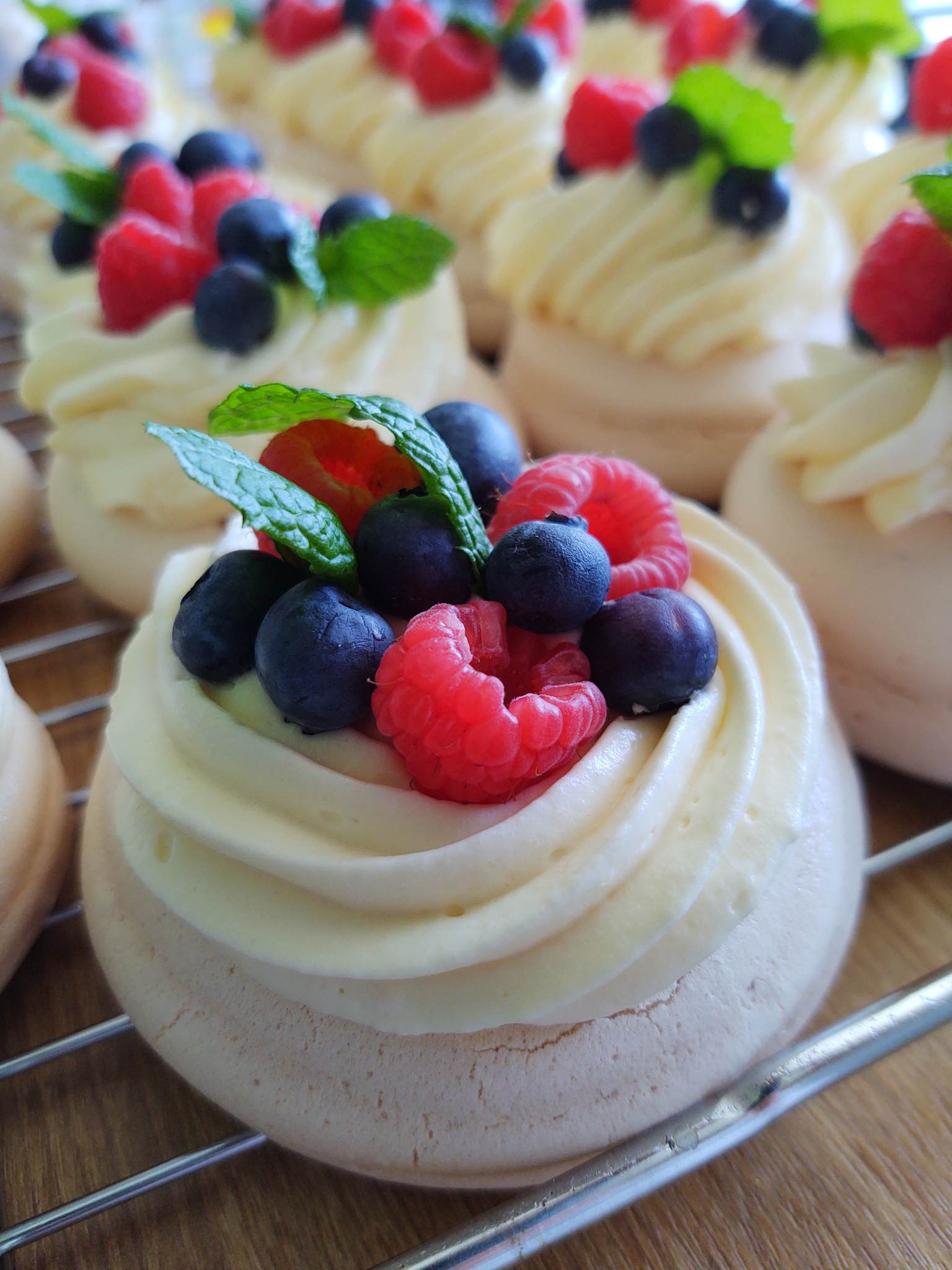 Beza Pavlova z kremem, malinami, borówkami i listkiem mięty na metalowej kratce.