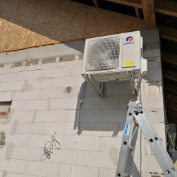 TOPSPLIT Łukasz Róż - Zewnętrzna jednostka klimatyzacji marki Gree zamontowana na ścianie budynku z białych bloczków, widoczna drabina Krause i żółty przewód wentylacyjny.