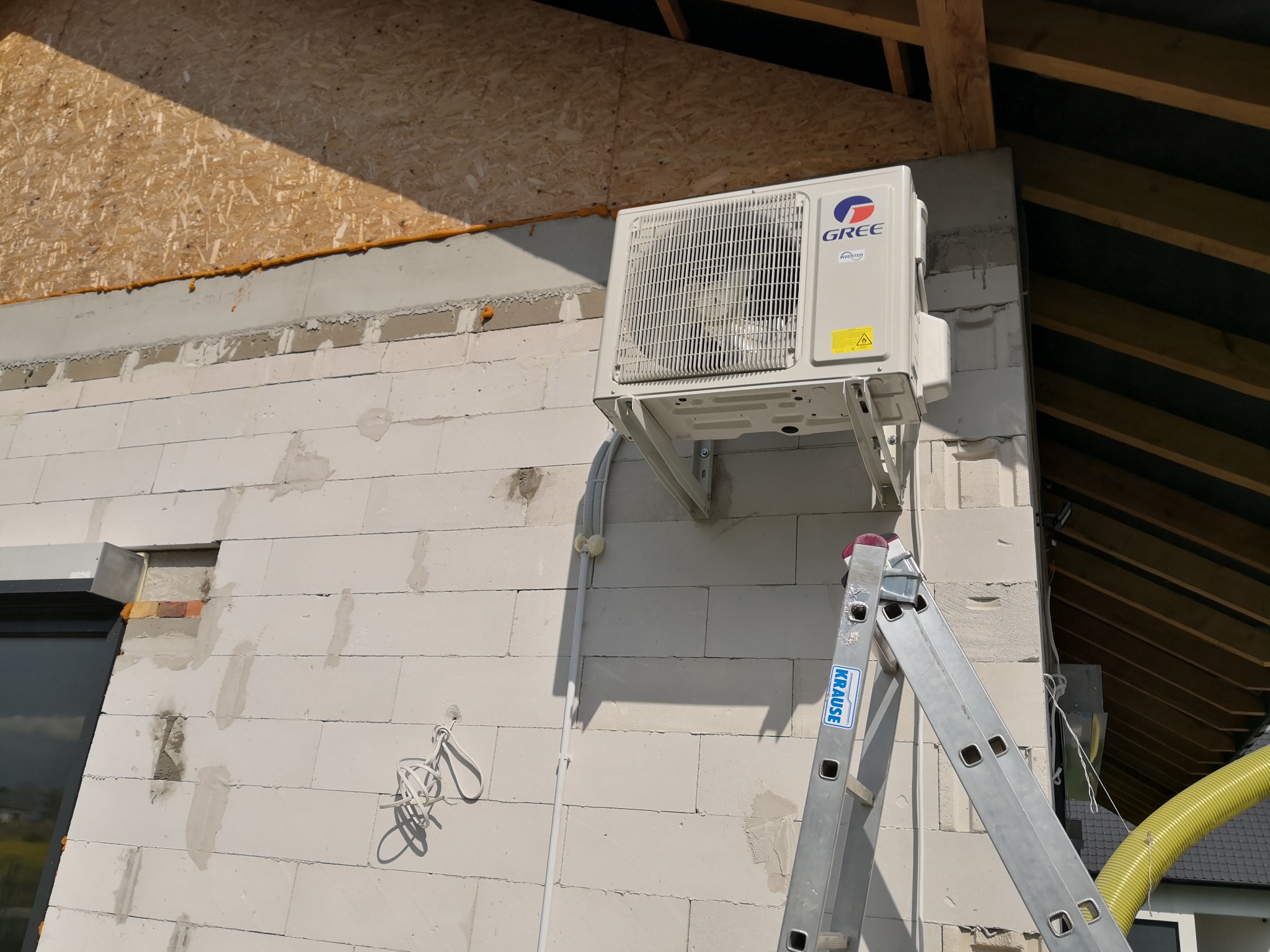 Zewnętrzna jednostka klimatyzacji marki Gree zamontowana na ścianie budynku z białych bloczków, widoczna drabina Krause i żółty przewód wentylacyjny.