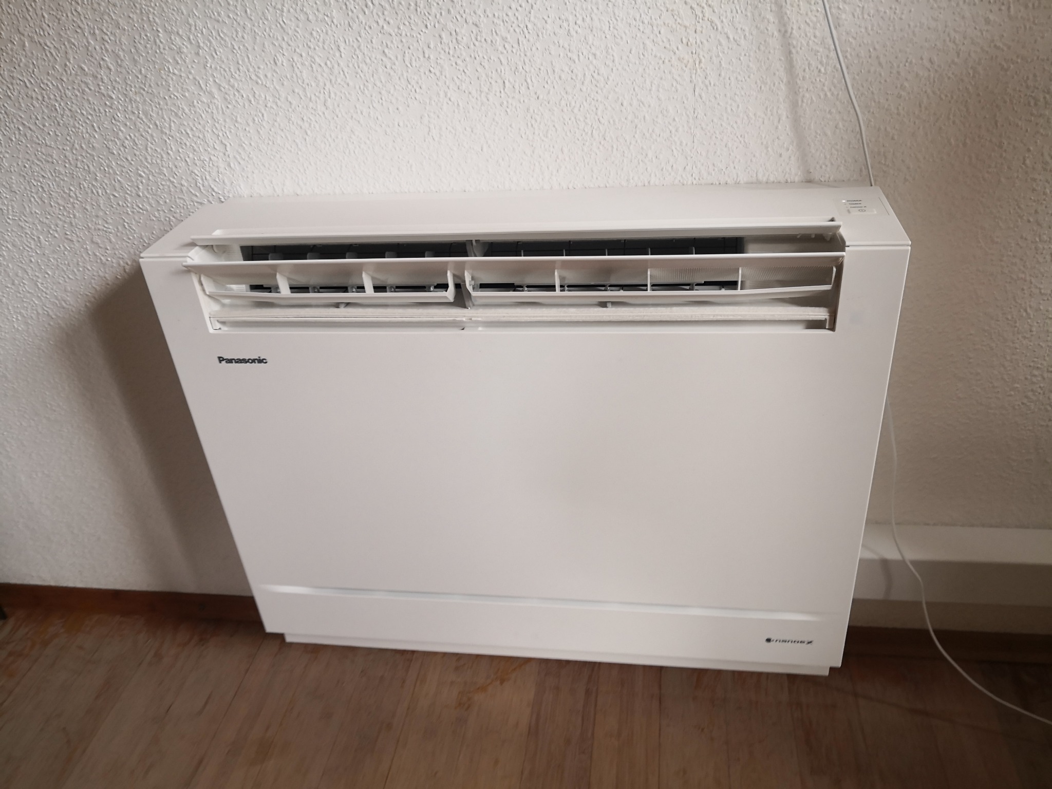 Biały, podłogowy klimatyzator Panasonic z otwartymi żaluzjami, ustawiony przy białej ścianie z widocznym przewodem zasilającym, na drewnianej podłodze.
