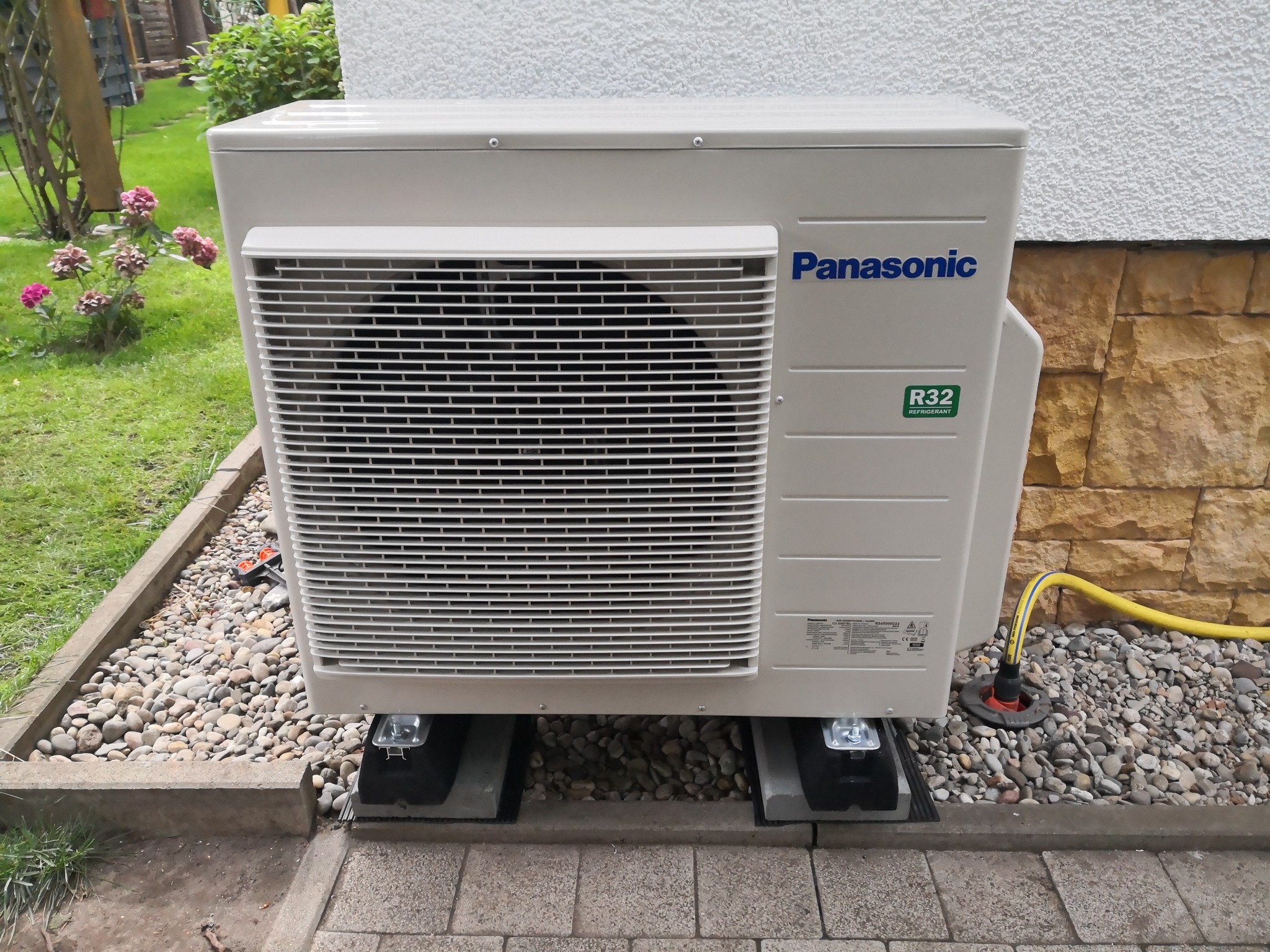 Zewnętrzna jednostka klimatyzacji Panasonic zamontowana na betonowych podstawach, otoczona kamieniami i kostką brukową, z widocznym przewodem odprowadzającym skropliny.