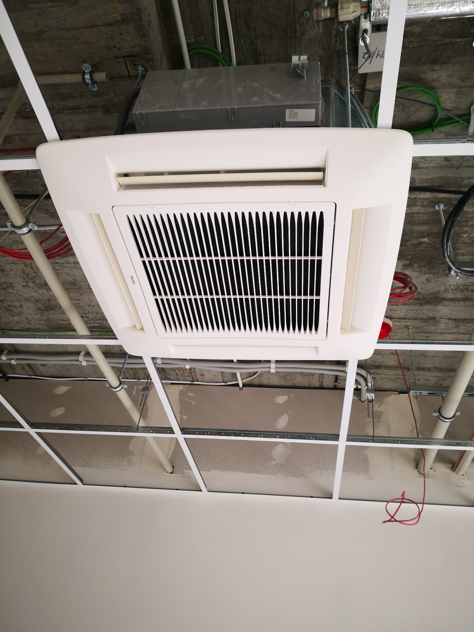 Klimatyzator kasetonowy zamontowany w suficie podwieszanym, widoczne rury i przewody instalacyjne.