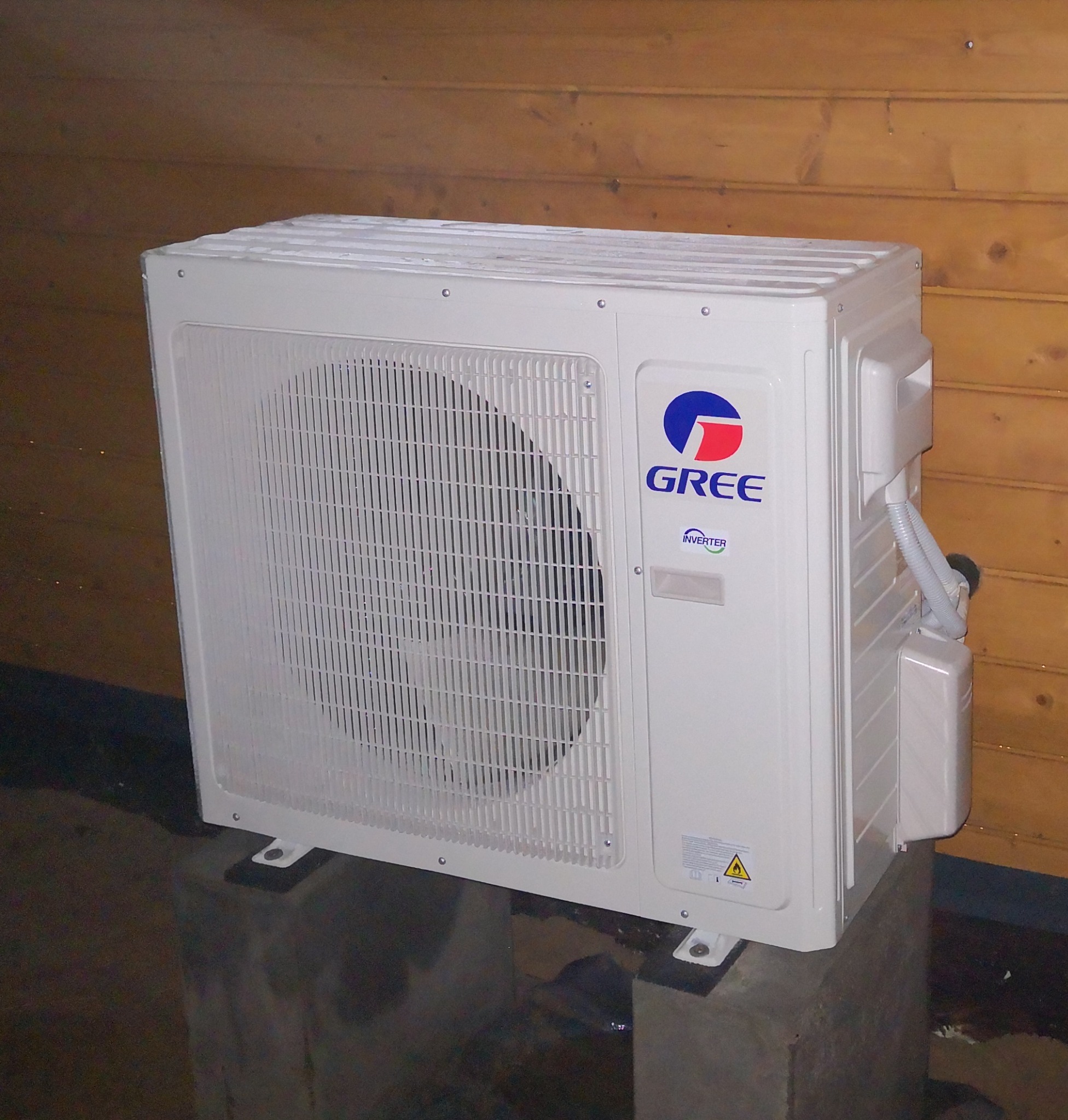 Zewnętrzna jednostka klimatyzacji marki Gree, zamontowana na betonowym fundamencie, z drewnianą ścianą w tle.