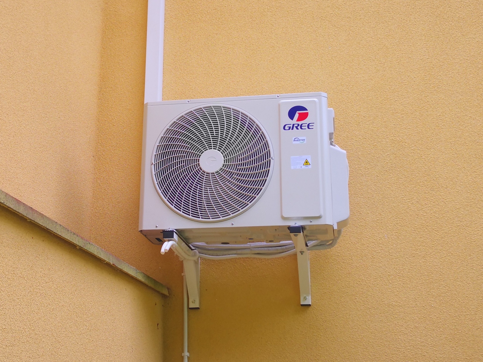 Zewnętrzna jednostka klimatyzacji Gree zamontowana na żółtej, teksturowanej ścianie budynku, widoczne białe rury odprowadzające skropliny i przewody.