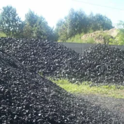 Góra czarnego węgla kamiennego na składzie, z widocznym betonowym ogrodzeniem w tle i zielenią drzew.