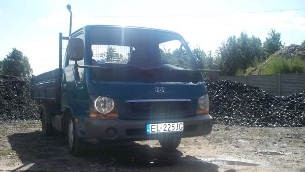 Niebieska ciężarówka Kia z polską tablicą rejestracyjną EL 225JG stoi na placu z pryzmą węgla w tle.