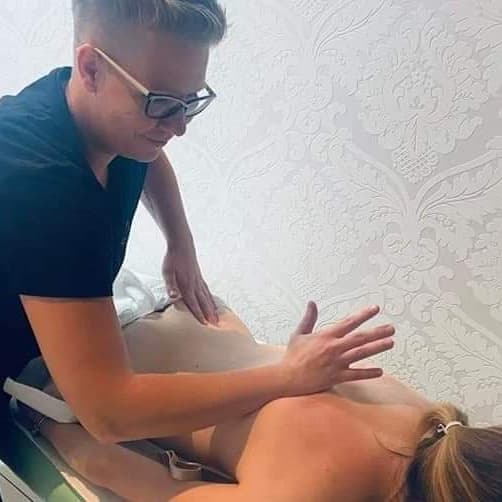 Terapeuta w okularach uciska dłonią dolną część pleców pacjentki leżącej na leżance, wzorzysta tapeta w tle.