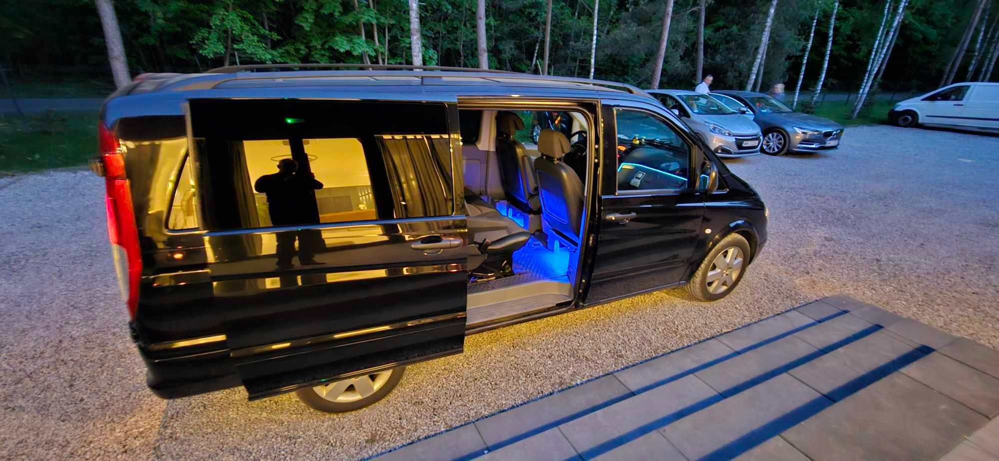 Czarny van z otwartymi drzwiami, podświetleniem LED wewnątrz i zasłonami w oknach, stojący na żwirowym parkingu.