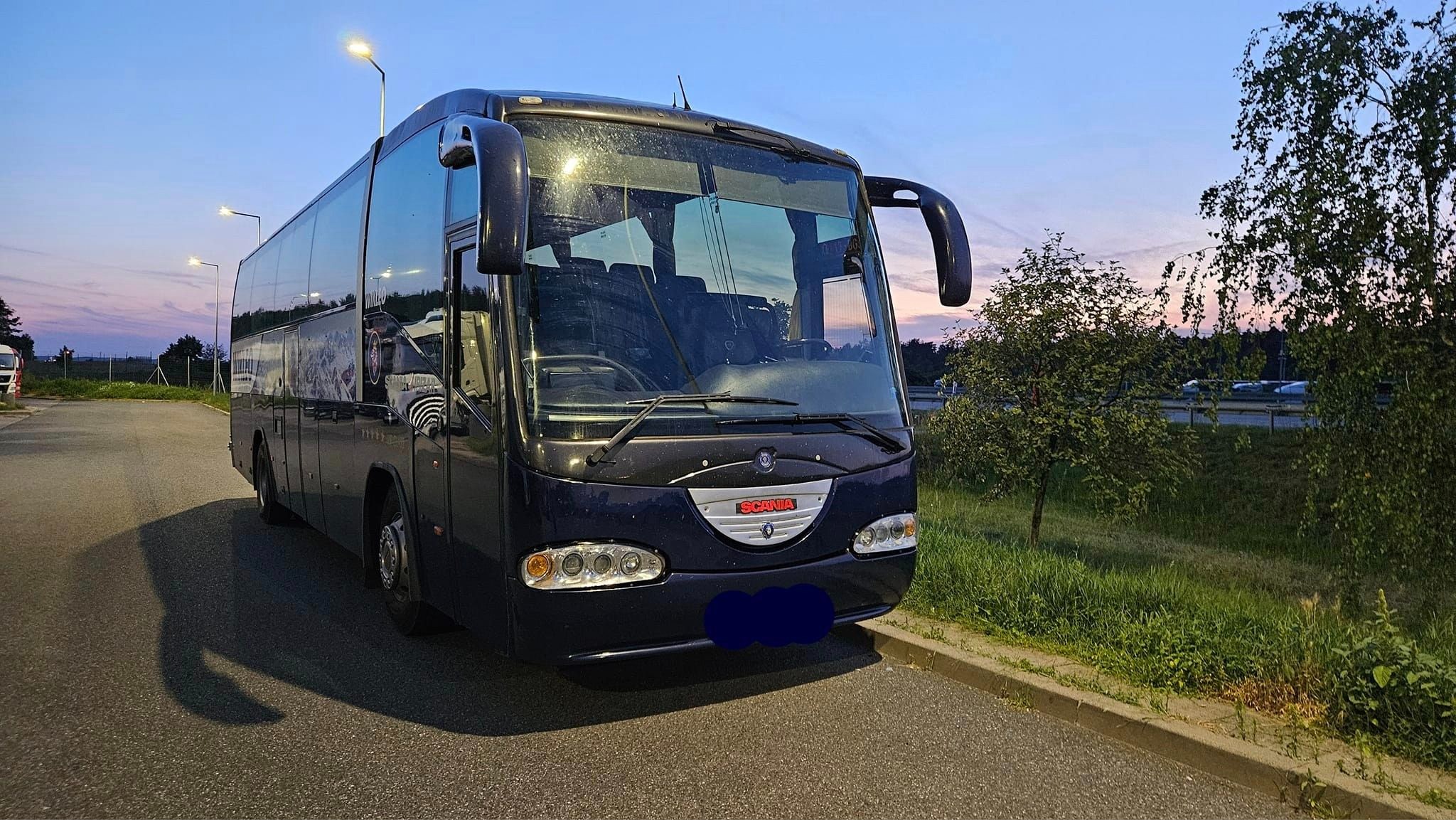 Czarny autokar Scania zaparkowany na parkingu o zmierzchu, widoczne odbicia w szybach i zielona roślinność w tle.