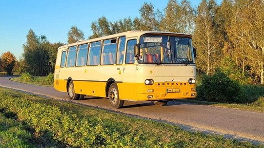 Żółto-biały autobus Autosan na asfaltowej drodze z zielonym poboczem i drzewami w tle, widoczne tablice rejestracyjne.