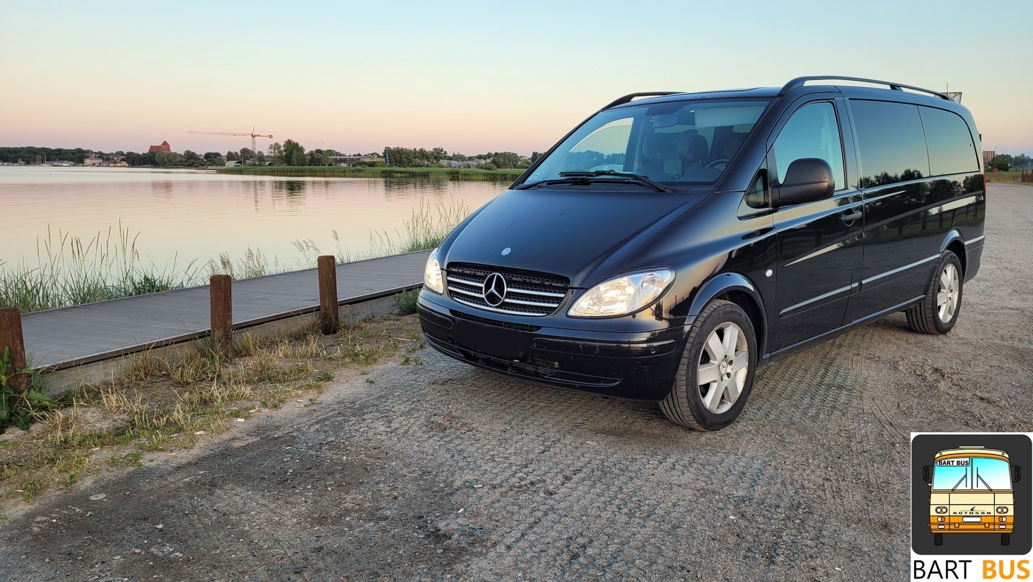 Czarny Mercedes Vito zaparkowany nad brzegiem jeziora o zachodzie słońca, w tle widoczny zamek i dźwig.
