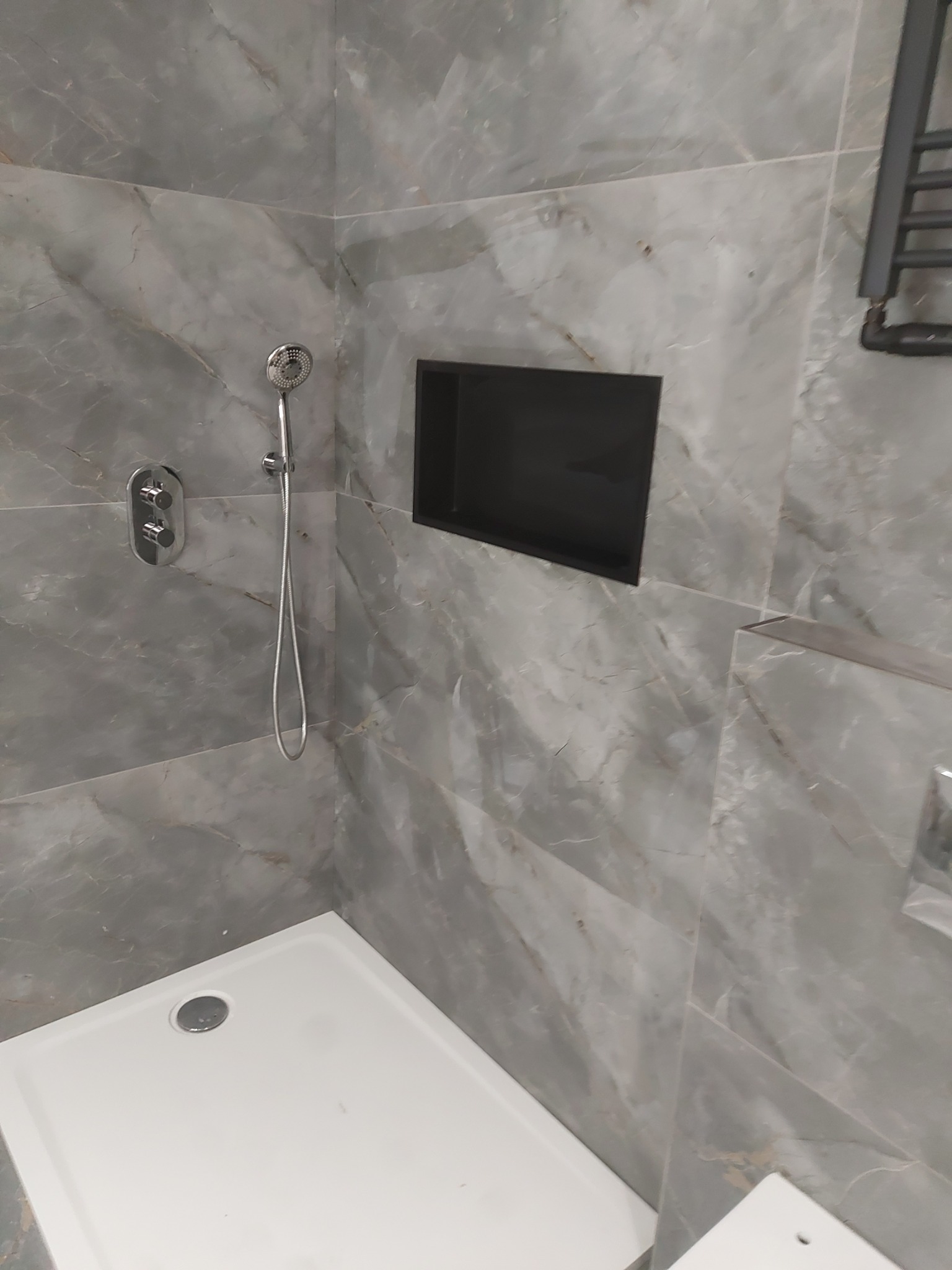 Wykończona kabina prysznicowa z szarymi płytkami imitującymi marmur, wnęką w ścianie i deszczownicą. Nowoczesny design, minimalistyczne wykończenie.