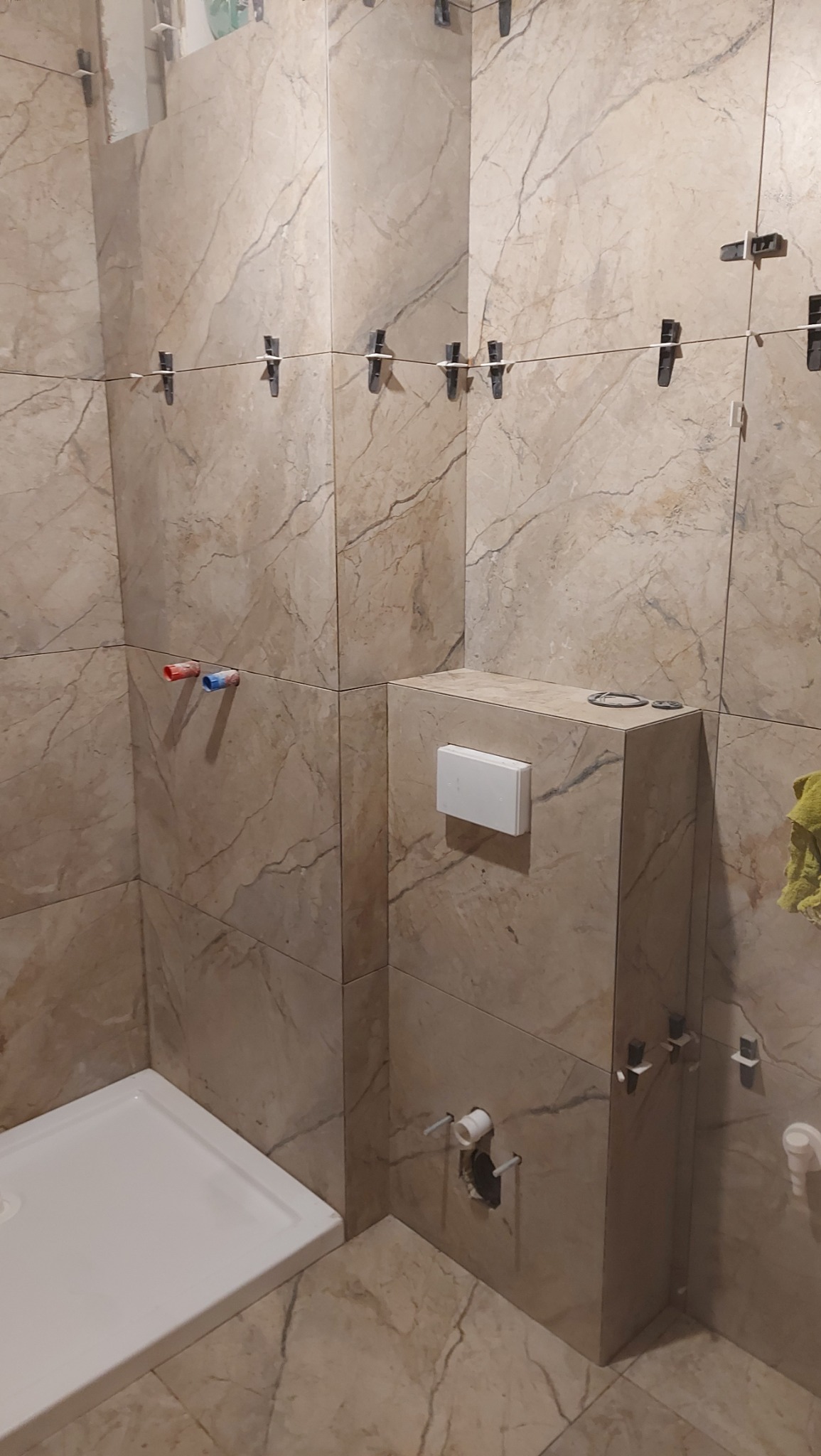 Łazienka w trakcie wykańczania: beżowe płytki imitujące marmur, system poziomowania płytek, zabudowa stelaża WC, brodzik. Widoczne przyłącza wodne.