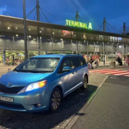 Błękitny minivan Toyota Sienna z polską tablicą rejestracyjną KTT-PG47 zaparkowany przed Terminalem A lotniska, pasy dla pieszych w tle.