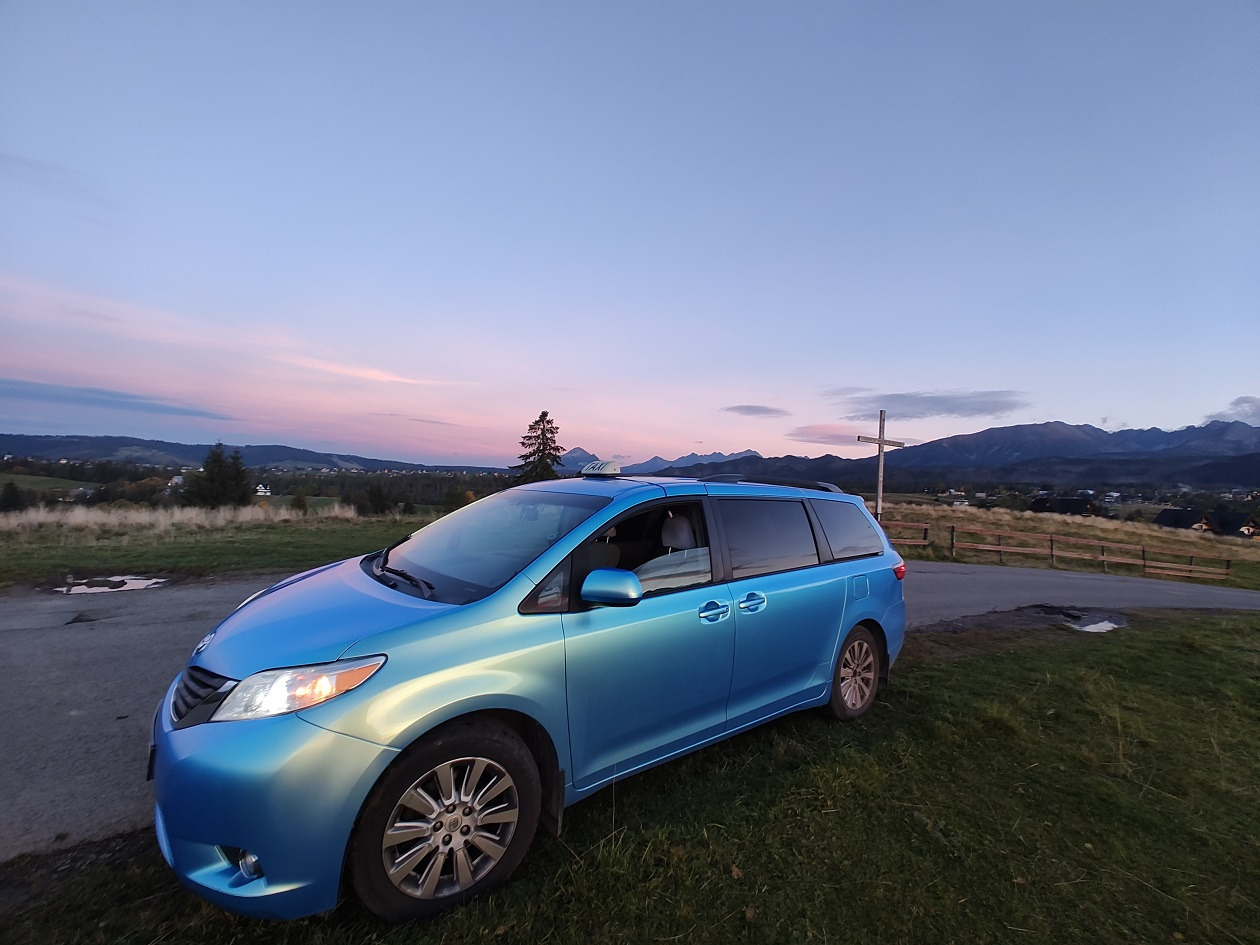 Błękitny minivan na tle górskiego krajobrazu o zmierzchu, widoczny krzyż na wzgórzu.