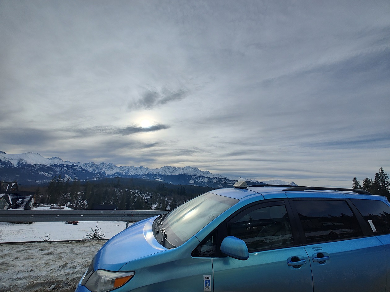 Błękitny van z bagażnikiem dachowym i oznaczeniem na drzwiach, na tle zimowego krajobrazu górskiego pod pochmurnym niebem.