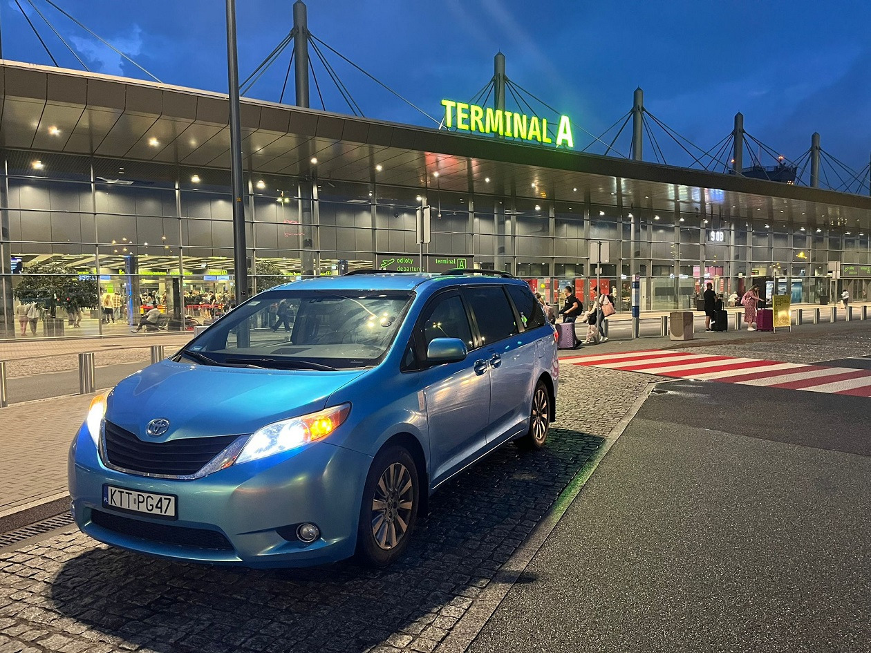 Błękitny minivan Toyota Sienna z polską tablicą rejestracyjną KTT-PG47 zaparkowany przed Terminalem A lotniska, pasy dla pieszych w tle.