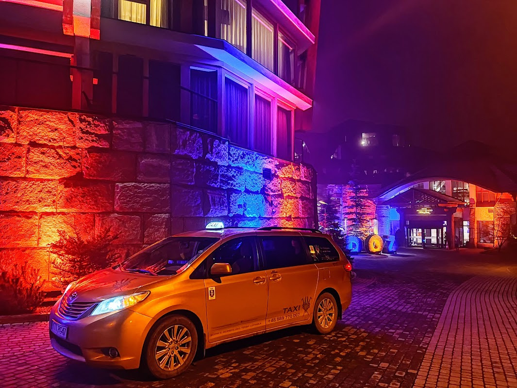 Złota taksówka minivan przed budynkiem oświetlonym czerwonym i niebieskim światłem, nocny widok.