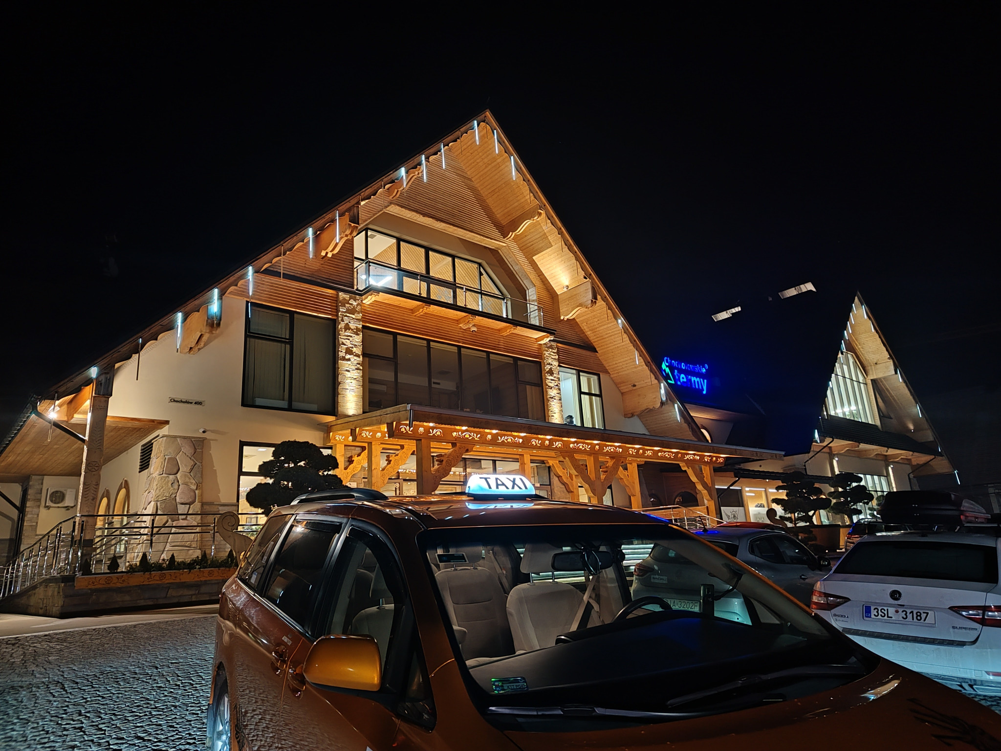 Pomarańczowy samochód taxi z podświetlonym napisem 'TAXI' na dachu, zaparkowany przed budynkiem w stylu góralskim z elementami kamiennymi i drewnianymi, oświetlonym nocą.