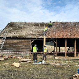 Gaja i Weles Spółka z Ograniczoną Odpowiedzialnością - Ekipa w kamizelkach odblaskowych demontuje dach starej, drewnianej stodoły z wykorzystaniem drabiny i deski, z zabezpieczeniem terenu taśmą ostrzegawczą.
