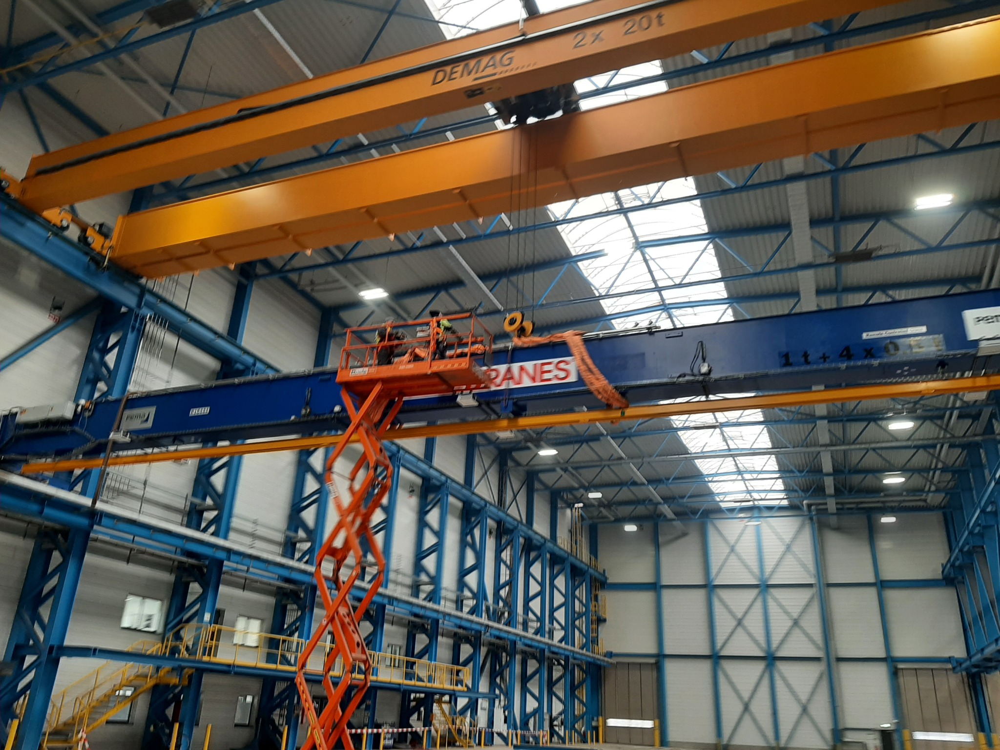 Montaż lub naprawa suwnicy w hali przemysłowej, widoczny podnośnik nożycowy z pracownikami w kaskach, żółta suwnica DEMAG 2x20t na niebieskiej konstrukcji hali.