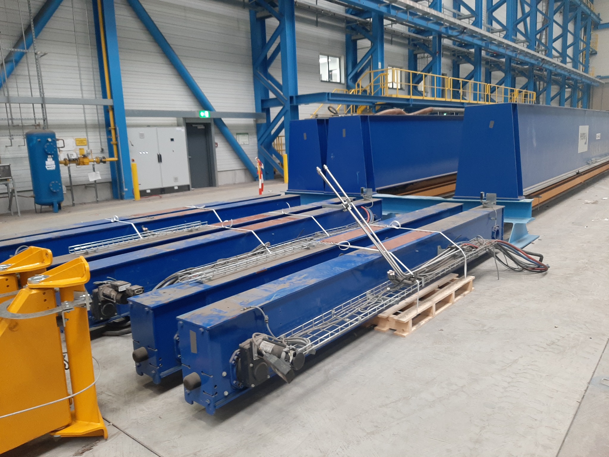 Niebieskie, stalowe elementy konstrukcyjne suwnicy pomostowej, z widocznymi silnikami i okablowaniem, ułożone na betonowej posadzce hali produkcyjnej, z żółtymi elementami z przodu i konstrukcją...