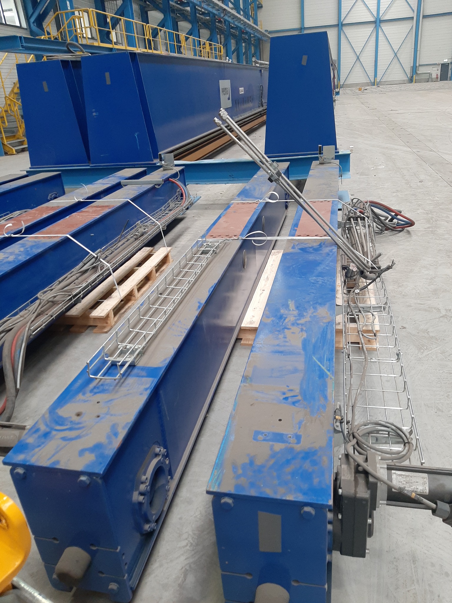 Elementy konstrukcyjne suwnicy pomalowane na niebiesko, widoczne kable i metalowe kosze na kable, całość w hali przemysłowej.