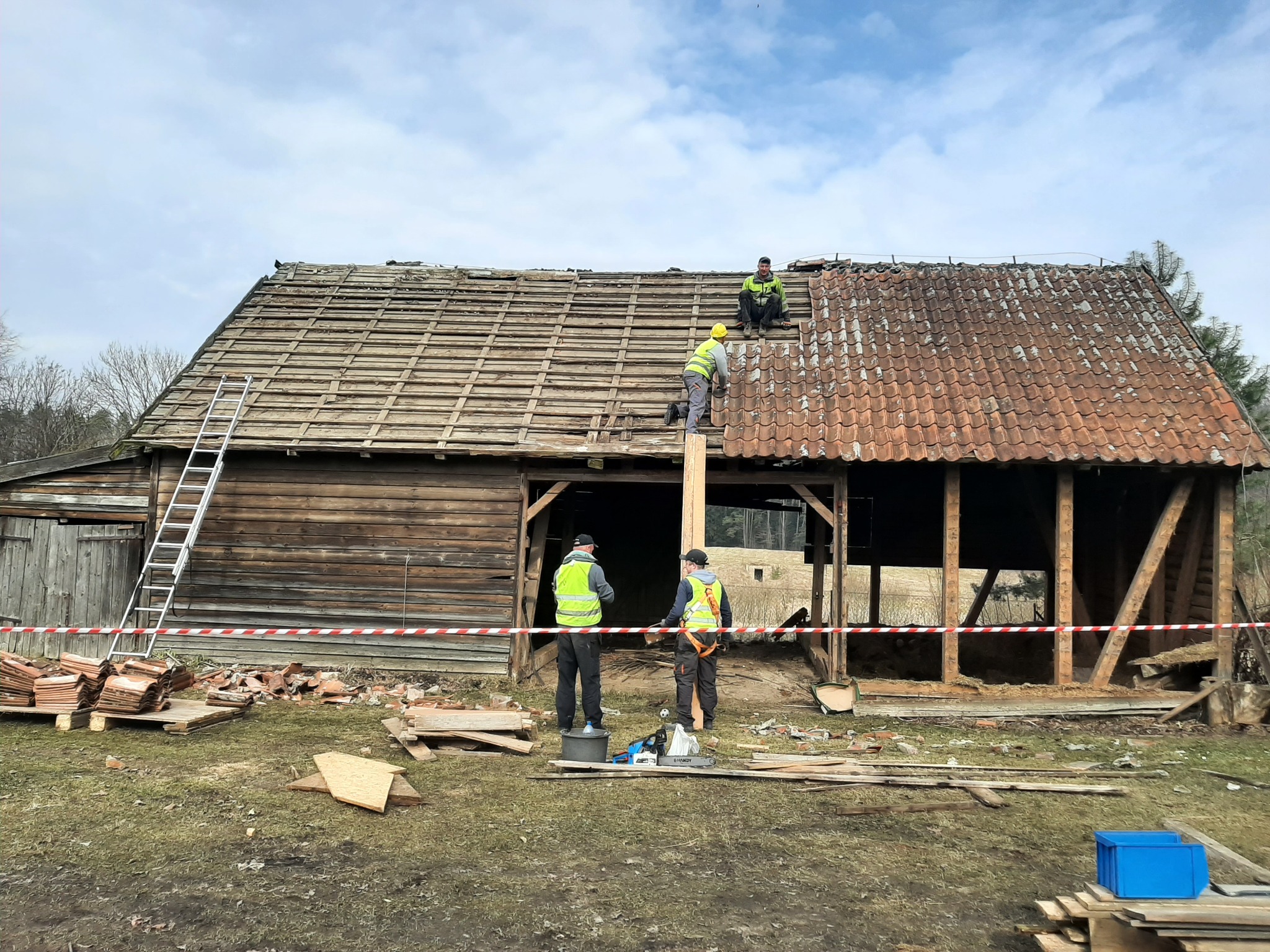 Ekipa w kamizelkach odblaskowych demontuje dach starej, drewnianej stodoły z wykorzystaniem drabiny i deski, z zabezpieczeniem terenu taśmą ostrzegawczą.
