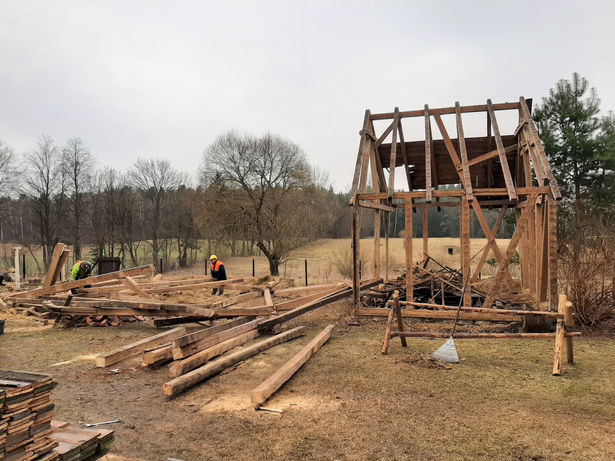 Demontaż drewnianej konstrukcji budynku na zewnątrz, widoczni dwaj pracownicy w odblaskowych kamizelkach, porozrzucane elementy konstrukcji, częściowo zdemontowany szkielet budynku.