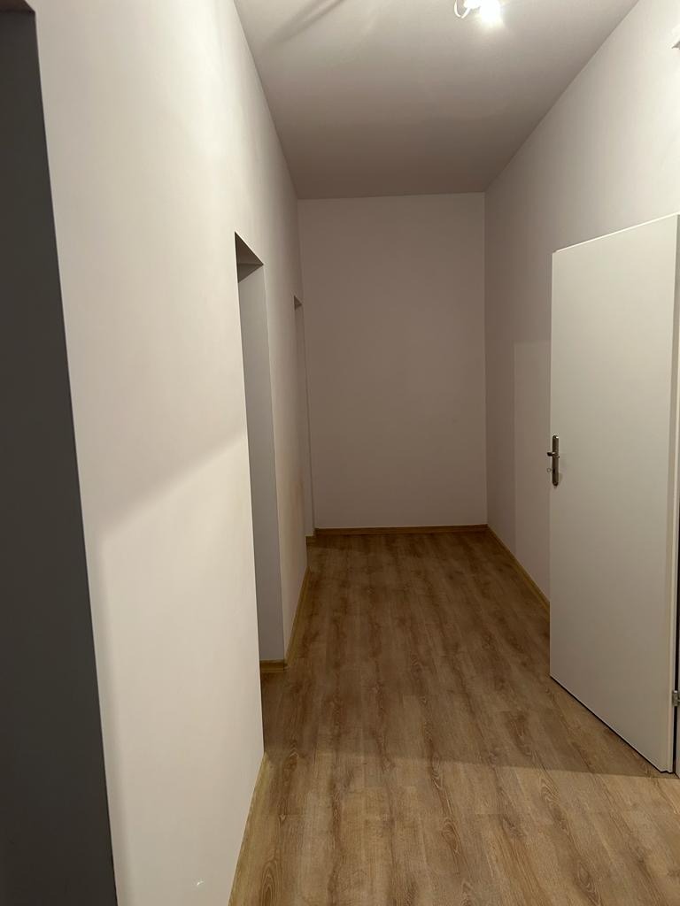 Korytarz po odświeżeniu: jasne ściany, panelowa podłoga imitująca drewno, białe drzwi z klamką, widoczne otwory drzwiowe.