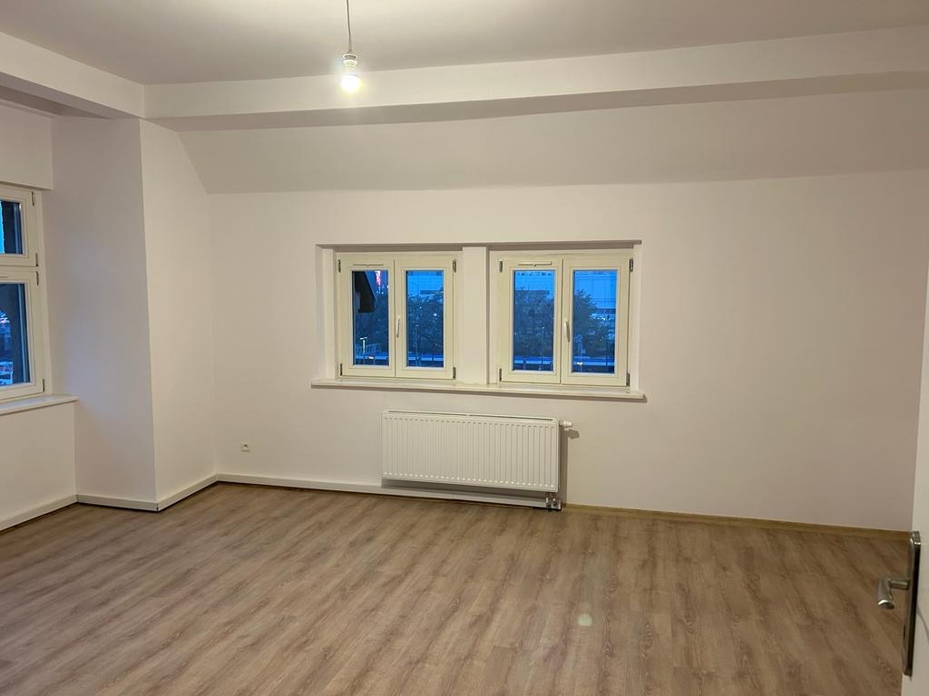 Pusty pokój po remoncie z panelami podłogowymi imitującymi drewno, białymi ścianami, grzejnikiem pod oknem z beżowymi ramami i pojedynczą żarówką sufitową.