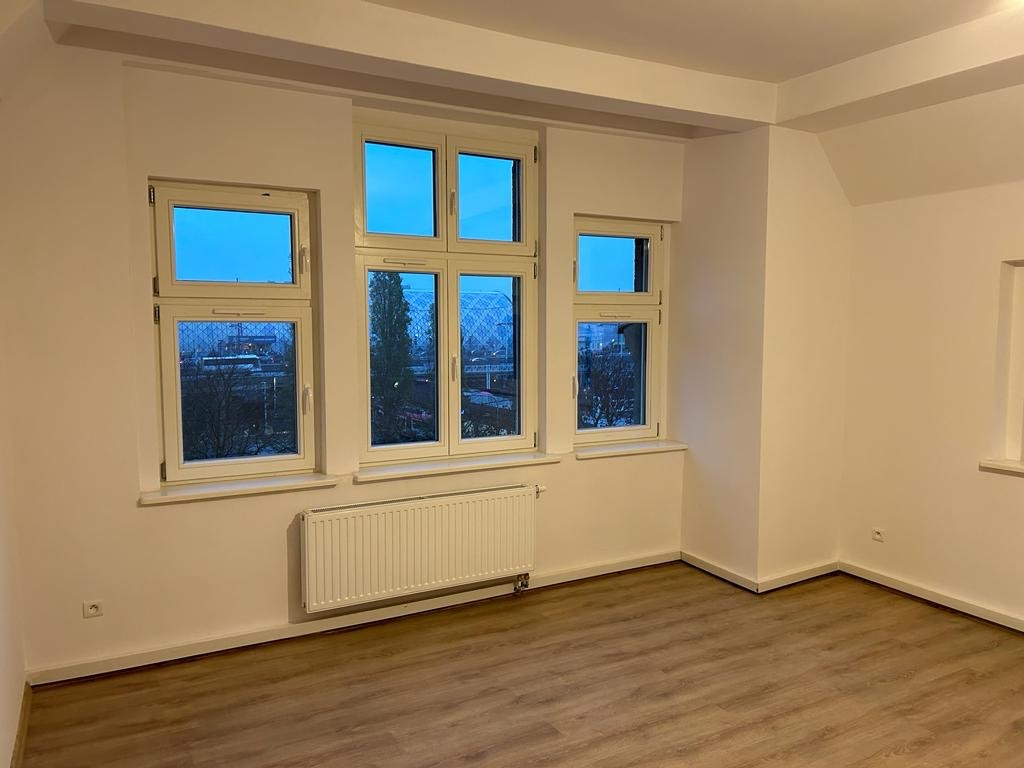 Puste, świeżo wykończone wnętrze z drewnopodobną podłogą, białymi ścianami, grzejnikiem i trzema oknami z widokiem na zewnątrz.