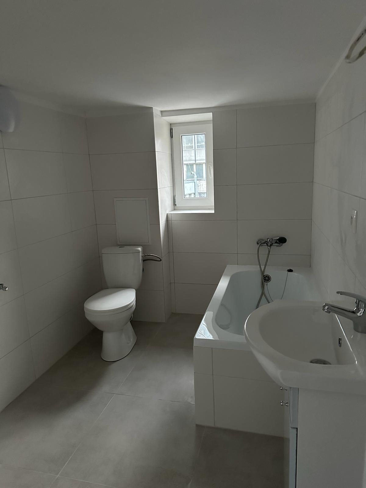 Jasna łazienka po remoncie z białymi płytkami, umywalką, wanną i toaletą, małe okno z widokiem na budynek.