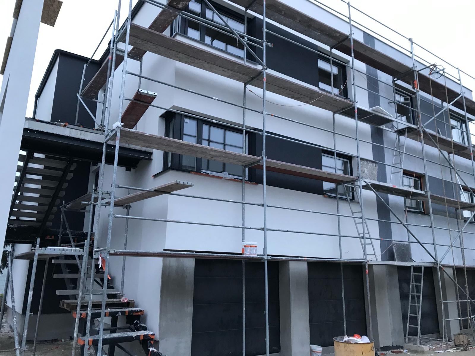 Dom w trakcie prac elewacyjnych, widoczne rusztowania, pomalowana część ściany na biało, czarne panele, ciemne okna, wiaderka z farbą na ziemi.