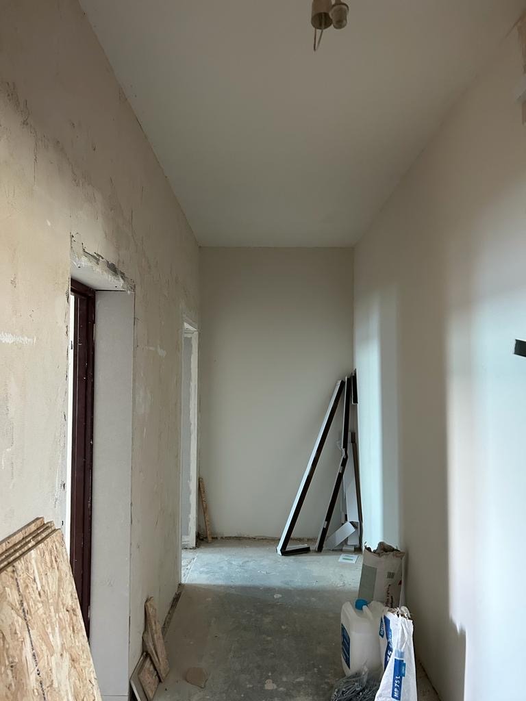 Korytarz w trakcie prac przygotowawczych: ściany z widocznymi śladami po kuciu, surowy tynk, widoczne płyty OSB, worki z zaprawą i profile aluminiowe oparte o ścianę.
