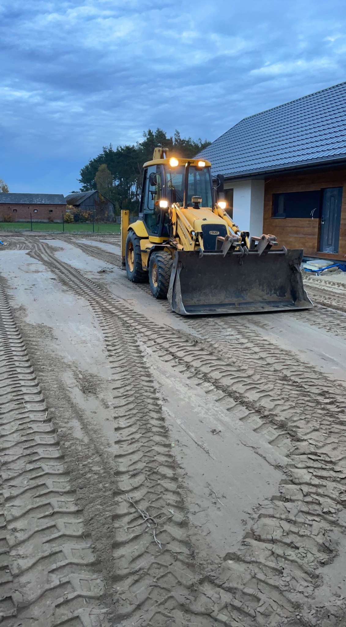 Żółta koparko-ładowarka na piaszczystym terenie z widocznymi śladami opon, w tle budynek mieszkalny z szarą dachówką i drewnianą elewacją, pochmurne niebo.