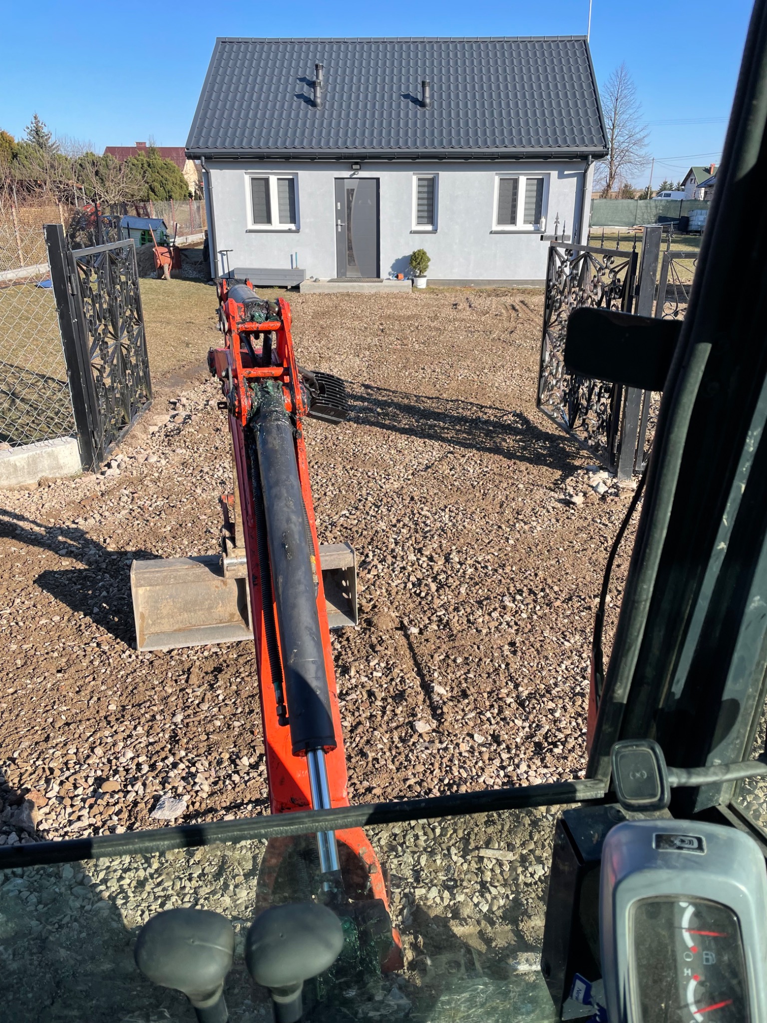 Widok z kabiny minikoparki na świeżo wyrównany teren przed domem z szarą elewacją i ozdobną bramą w słoneczny dzień.